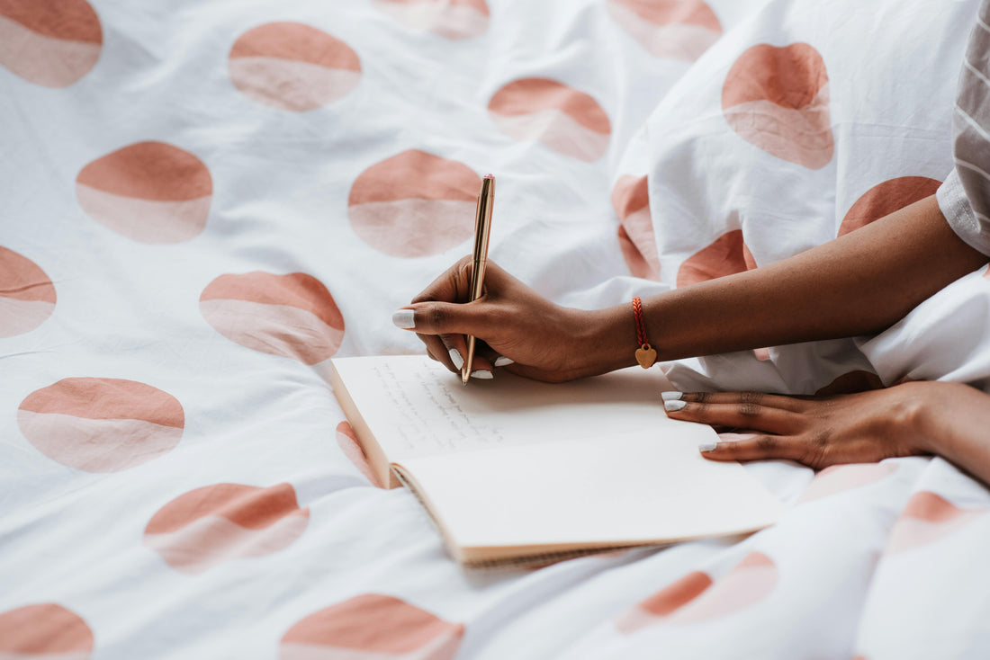 Woman journaling in bed with pink, polka-dotted sheets, gold pen in hand.
