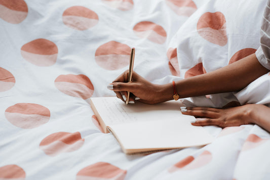 Woman journaling in bed with pink, polka-dotted sheets, gold pen in hand.